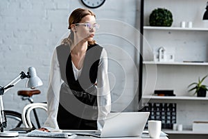 Businesswoman in formal wear and glasses standing near computer desk at workplace and