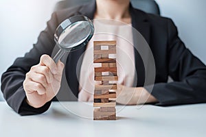 Businesswoman hand holding glass magnifying over wooden blocks tower. Business analysis, Risk Management, Study, opportunities and