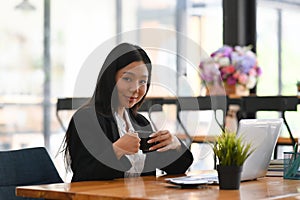 Businesswoman holding coffee cup and sitting in front of laptop computer in office.