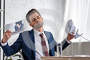Businesswoman holding electric ventilator and waving business newspaper while suffering from heat in office