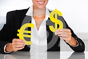 Businesswoman Holding Euro And Dollar Sign At Desk
