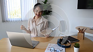 Businesswoman holding file folder and celebrating her success at office desk.