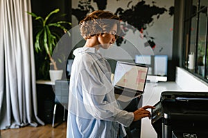 Businesswoman holding laptop and pressing button on panel of printer in office