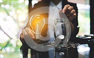 Businesswoman holding a light bulb over coins stack on the table while putting coin into a glass jar