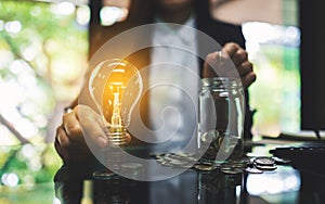 Businesswoman holding a light bulb over coins stack on the table while putting coin into a glass jar