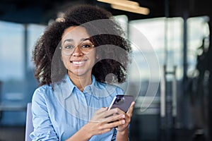 Businesswoman inside office at workplace close up smiling and looking at camera, office worker holding phone in hands