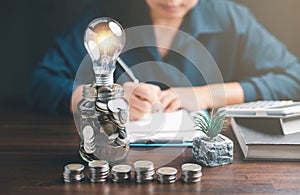 Businesswoman with lightbulb on money jar and using calculator to calculate and money stack. Save energy and money with accounting