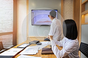 Businesswoman listening to businessman present the monthly college budget in front of TV during meeting