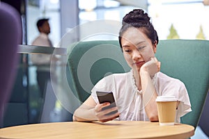Businesswoman With Mobile Phone Working At Table In Breakout Area Of Office Building 
