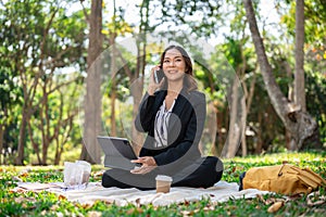 A businesswoman is multitasking, talking on the phone and working on her digital tablet in a park