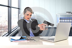 Businesswoman pointing gun to computer laptop sitting at desk desperate and stressed