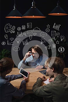 Businesswoman showing something on tablet to her partners and sitting at table with hand-drawn business icons