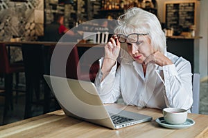 Businesswoman sits at table in front of laptop and looks closely at monitor, raising her glasses. Education for adults