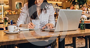 Businesswoman is sitting in cafe at table in front of laptop, making notes in notebook, working.Student learning online.
