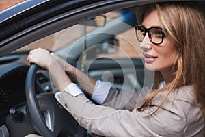 Businesswoman sitting in her car. Blonde with long hair and in glasses.