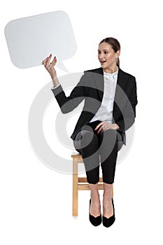 Businesswoman sitting and holding speech bubble and looking aside fascinated