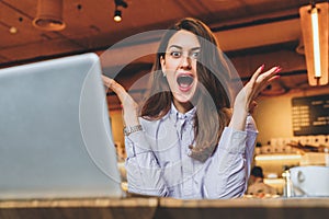 Businesswoman is sitting at table in front of laptop and opening her mouth,raising her hands in surprise looks at screen