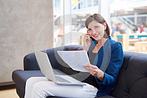 Businesswoman smiling with paperwork, phone and laptop on couch