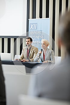Businesswoman speaking through microphone while sitting with colleague in seminar hall