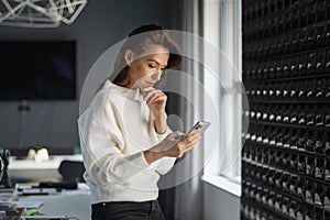 Businesswoman standing at the office by the window and using mobile phone