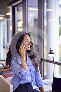 Businesswoman Standing By Partition In Open Plan Office Making Call On Mobile Phone