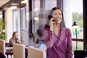 Businesswoman Standing By Partition In Open Plan Office Making Call On Mobile Phone