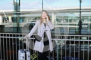 Businesswoman standing with valise and talking by smartpone near airport.