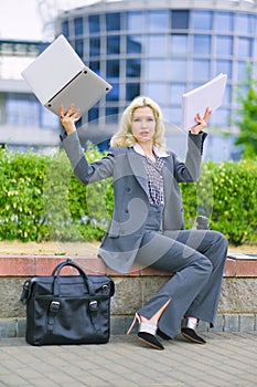 A businesswoman on the street holds a laptop and paper in her hand. The woman has her hands in the air