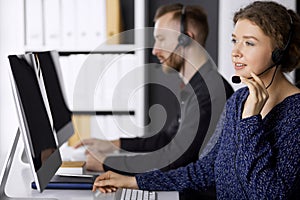 Businesswoman talking by headset while sitting with red-bearded colleague in modern office. Telemarketing and customer