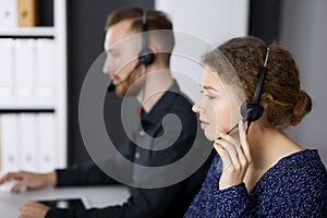 Businesswoman talking by headset while sitting with red-bearded colleague in modern office. Telemarketing and customer