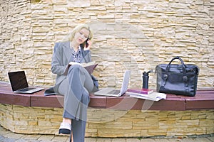 A businesswoman is talking on the phone. A woman sits on a bench with a laptop and notepad.