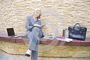 A businesswoman is talking on the phone. A woman sits on a bench with a laptop and notepad.