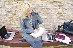 A businesswoman is talking on the phone. A woman sits on a bench with a laptop and notepad.