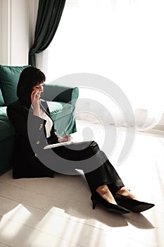 Businesswoman is talking on a smartphone. Tired brunette woman is sitting close to green sofa in a business black suit