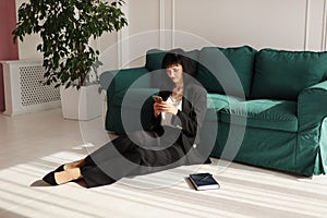 Businesswoman is talking on a smartphone. Tired brunette woman is sitting close to green sofa in a business black suit