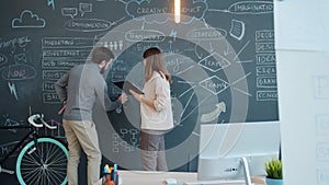 Businesswoman talking to male colleague who is writing information on chalkboard in office