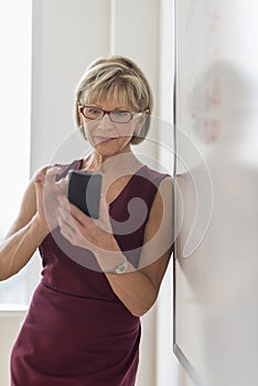 Businesswoman Text Messaging On Smart Phone While Leaning On Whiteboard