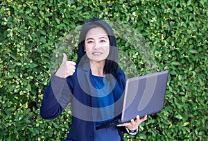 Businesswoman thumps up and holding laptop computer at green lea