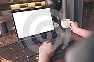 A businesswoman using laptop with blank white desktop screen while drinking hot coffee on wooden table in cafe