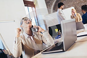 Businesswoman using laptop and phone with team discussing project in the background