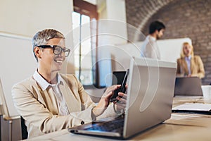 Businesswoman using laptop and phone with team discussing project in the background