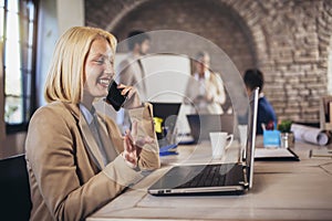 Businesswoman using laptop and phone with team discussing project in the background