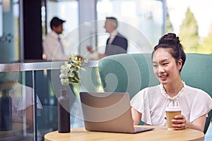 Businesswoman Using Laptop Working At Table In Breakout Seating Area Of Office Building 