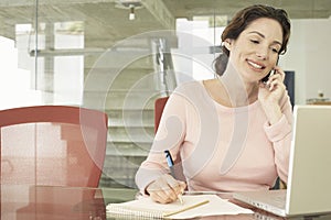 Businesswoman Using Mobile Phone While Writing On Notepad