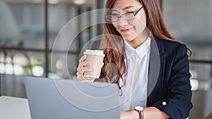 Businesswoman using and typing on laptop while working and drinking coffee in office