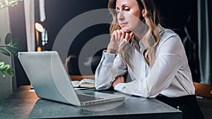 Businesswoman in white shirt is sitting in office at table in front of computer and pensively looks at screen of laptop