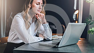 Businesswoman in white shirt is sitting in office at table in front of computer and pensively looks at screen of laptop