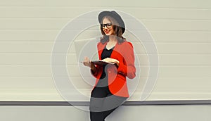 Businesswoman working with laptop wearing red blazer jacket, eyeglasses, round hat on gray background, happy elegant woman holds
