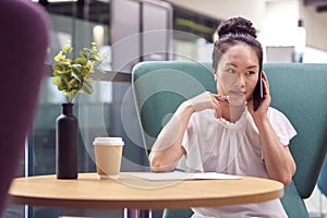 Businesswoman Working At Table In Breakout Seating Area Of Office Building Talking On Mobile Phone