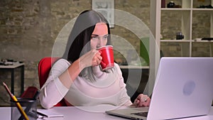 Businesswoman works in the office in front of the laptop, drinks coffee and simultaneously watch the screen